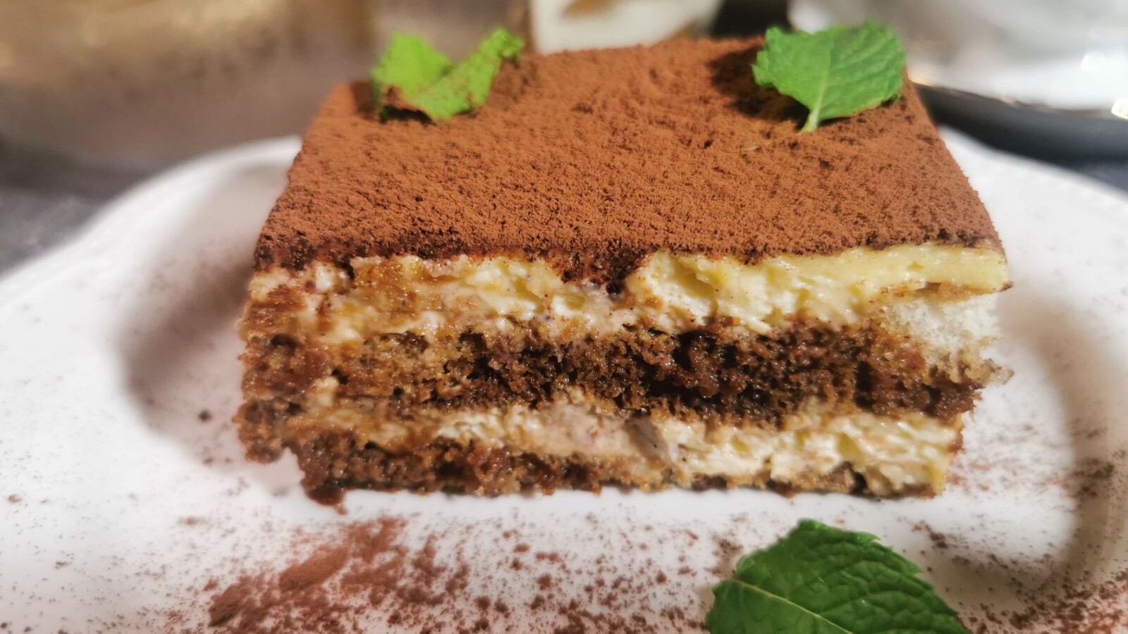Presjek tiramisua s vidljivim slojevima kreme i piškota, posut kakaom i dekoriran mentom.