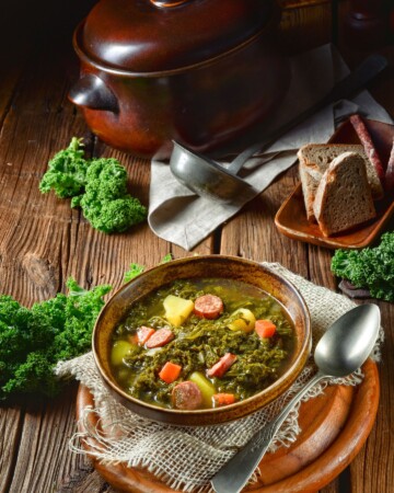 Zdjela toplog kelj variva s krumpirom, mrkvom i kuhanom kobasicom, poslužena na rustikalnom drvenom stolu uz svježe listove kelja, kruh i žlicu.