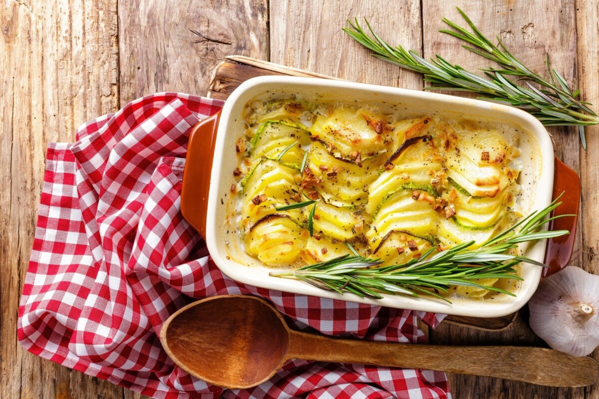 Gratinirani krumpir u keramičkoj posudi, poslužen na drvenoj površini uz crveno-bijelu kuhinjsku krpu, drvenu žlicu, češnjak i grančice ružmarina.